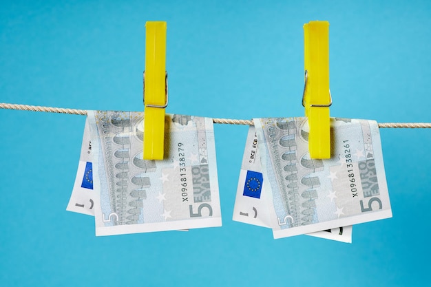 Five euro banknotes fastened with clothespins are dried on a clothesline on blue background