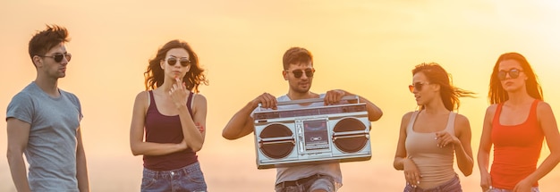 The five happy people stand with a boom box on the sunset background