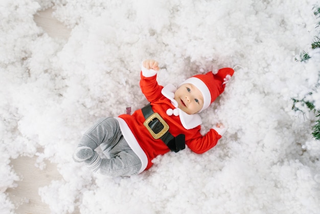 A five-month-old little child in a Santa Claus suit is lying in artificial snow on his back