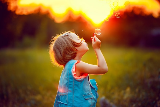 Five years old caucasian child girl blowing soap bubbles outdoor at sunset .