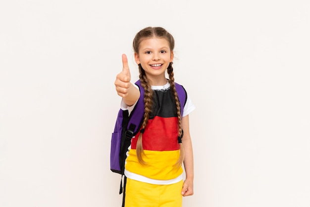 The flag of Germany on the Tshirt of a little girl A beautiful little girl with pigtails and a satchel behind her back shows a thumbs up on a white isolated background Language courses for children