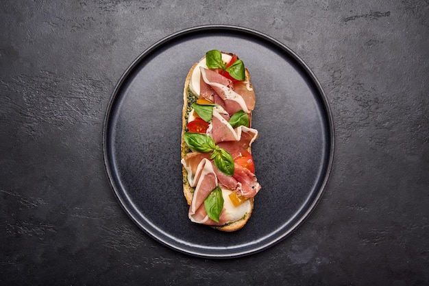 Flat lay bruschetta with mozzarella parma ham and pesto on black plate on graphite background