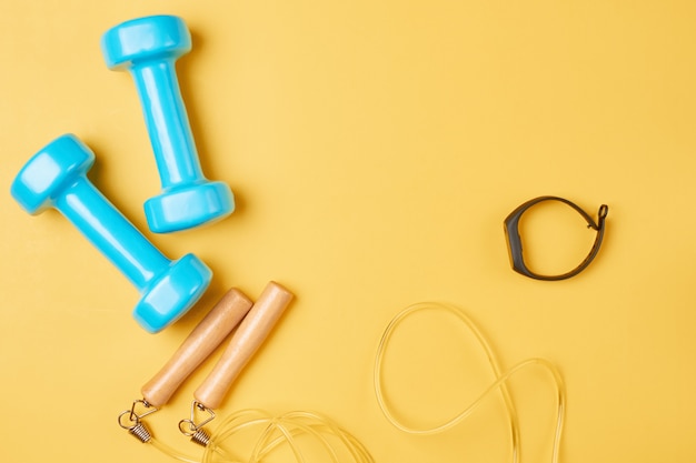 Photo flat lay composition of blue dumbbells, jump rope and fitness tracker on a yellow background