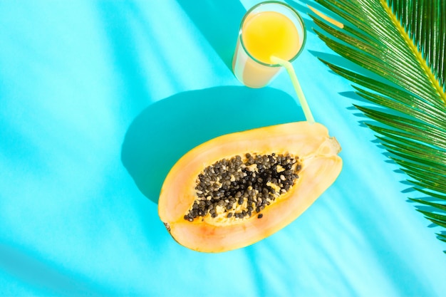 Flat Lay Composition Tall Glass with  Fruit Juice Halved Papaya, Palm Leaf on Blue Background.