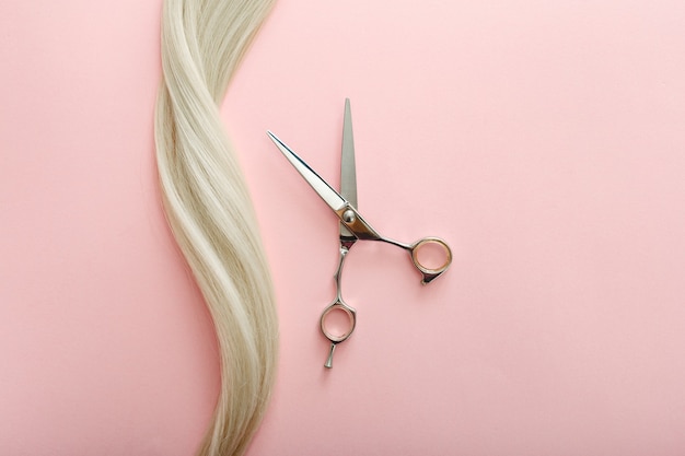 Flat lay composition with Hairdresser tools - scissors and strand of blonde hair on pink color background with copy space for text. Hairdresser service. Beauty salon service.