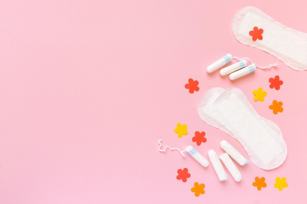 Flat lay of female menstrual supplies Tampons and pads with paper flowers on pink background space for text