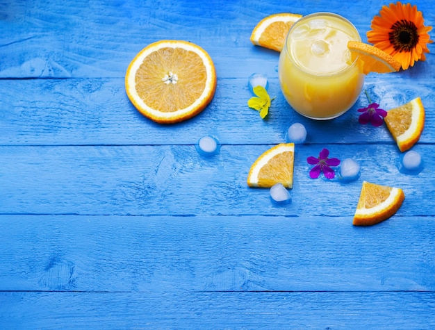 Flat lay of fresh orange juice in a glass with slices of ripe oranges ice cubes and flowers on the blue wooden table