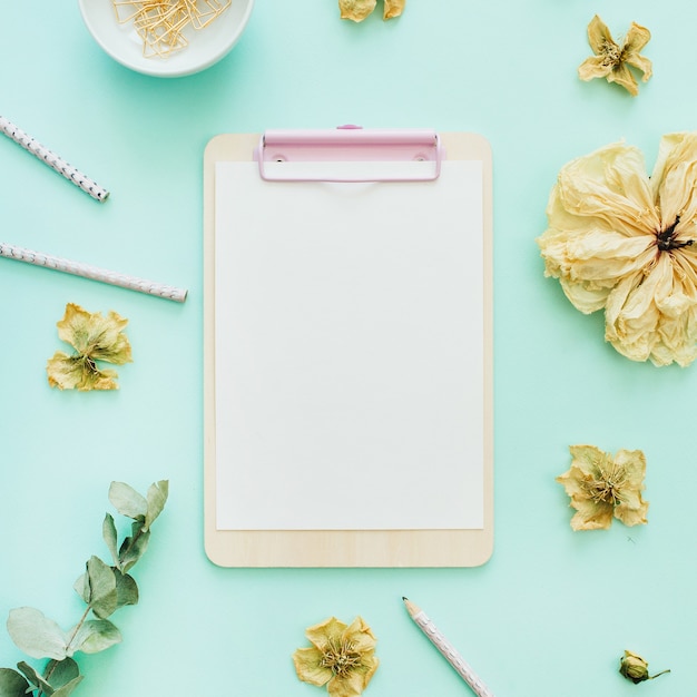 Flat lay home office desk workspace with clipboard, flowers and branches on blue background. Top view