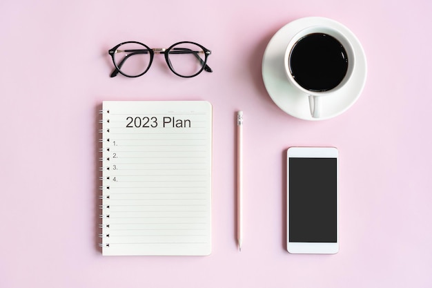 Flat lay of mobile phone a cup of coffee glasses and notepad of businesswoman on pink table in office 2023 Plan concept