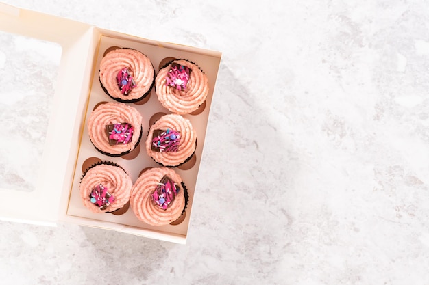 Flat lay Packaging freshly baked chocolate strawberry cupcakes garnished with gourmet mini pink chocolates into a white paper cupcake box