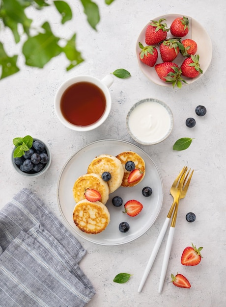 Flat lay pancakes with cottage cheese with fresh blerries and sour cream on a gray background with a cup of tea The concept of a healthy and delicious morning breakfast Top view