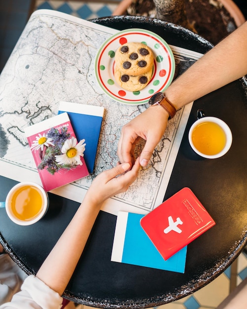 Flat lay photo of man and woman hands touching each other. Pair planning vacation. On the black table different travel guides, map, cups with tea, cookies and passport. Wanderlust concept.