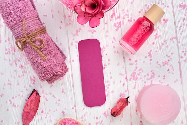 Flat lay pink spa accessories on white wooden table. Rolled towel with lotion, candle etc.