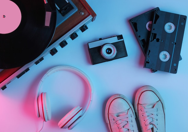 Flat lay retro 80s pop culture objects. Vinyl player, headphones, video tapes, film camera, sneakers with gradient neon blue-pink light. Retro wave. Top view