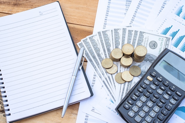 Flat Lay of stack of coins on dollar banknote , calculator, notebook, pen, blue pastel chart paper on the wooden table. Business, finance, marketing, e-commerce concept.