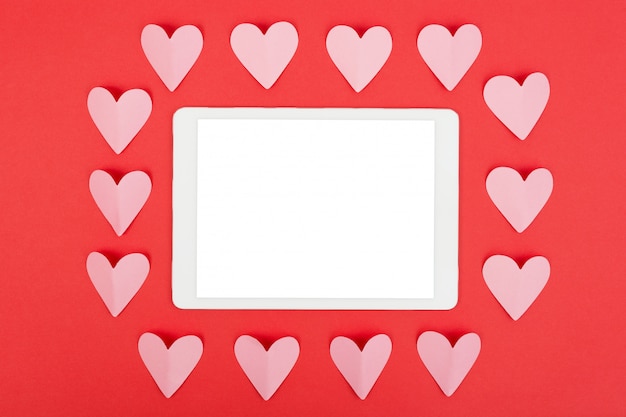 Flat lay of tablet computer with blank screen on red background with paper hearts