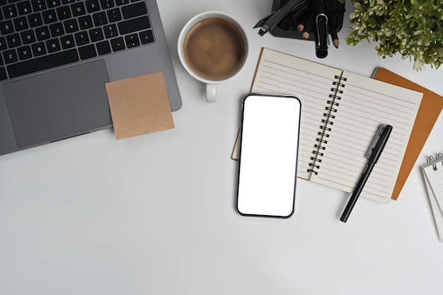 Flat lay top view of smart phone with empty screen notepad laptop and coffee cup on white working desk