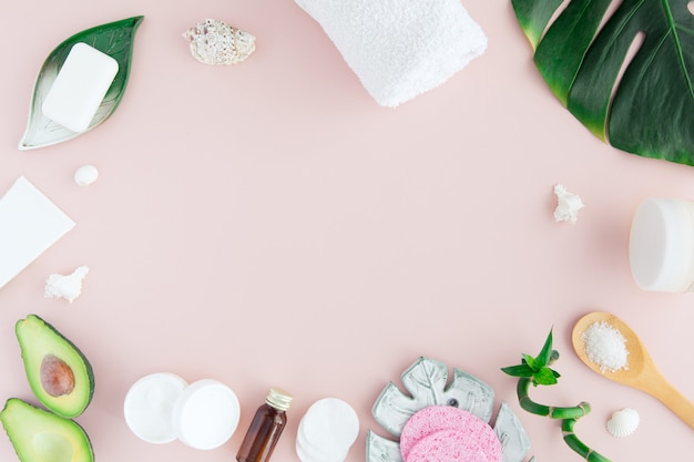 Flat lay and top view of white towel, jar of cream, green leaves and bamboo, avocado on pastel pink