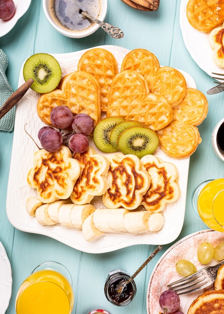 Flat lay with healthy breakfast with fresh hot waffles hearts, pancakes flowers with berry jam and fruits on turquoise surface, top view, flat lay. Food concept