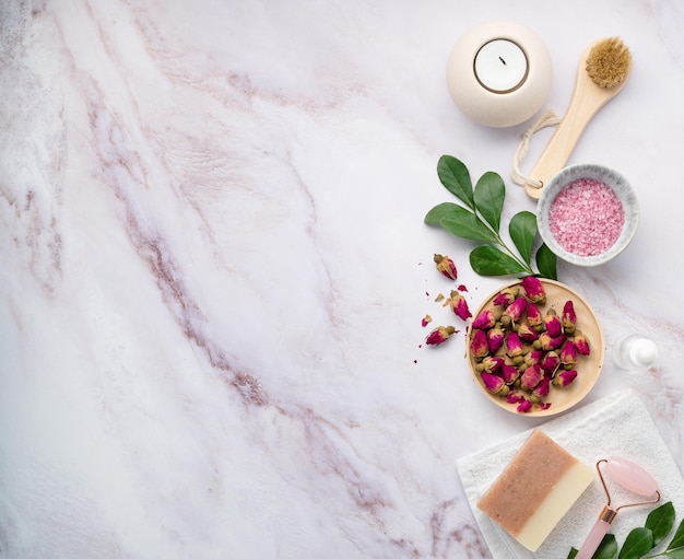 Flat lay with organic sea salt with dried rose flowers soap candle brush and gua sha on a pink marble background The concept of a natural spa product Top view and copy space