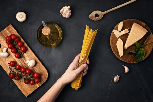 Flat lay of woman hand holding spaghetti and ingredients for cooking italian pasta. Spaghetti, tomatoes, oil, garlic, parmesan. Top view of traditional italian cusine concept