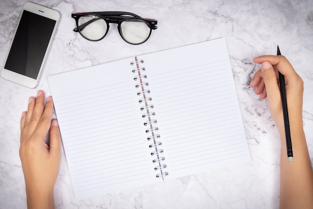 Flat lay of woman hand writing in a blank white page notebook on white marble desk