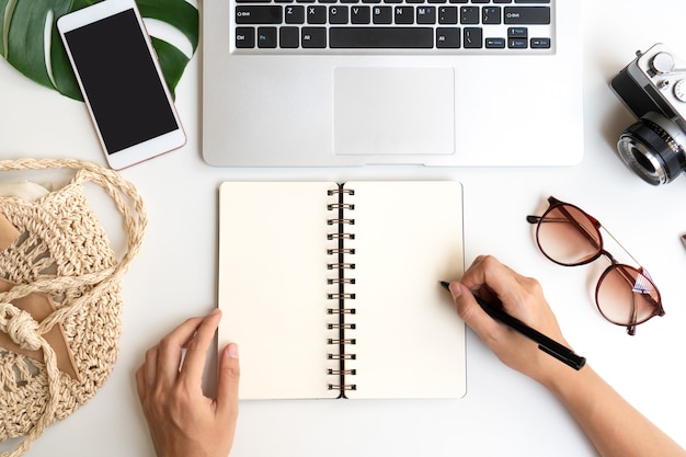 Flat lay of woman hands writing on notebook with travel accessories and laptop on desk