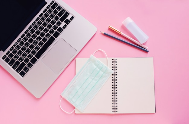 Flat lay of workspace desk with laptop, blank notebook, medical masks and alcohol gel on bright pink