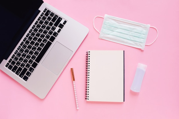 Flat lay of workspace desk with laptop, blank notebook, medical masks and alcohol gel on bright pink