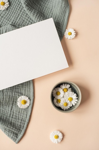 Flatlay aesthetic Parisian styled invitation template Blank paper sheet card with mockup copy space chamomile flower buds and aquamarine muslin cloth on neutral beige background Flat lay top view