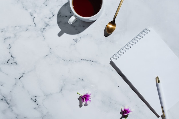 Flatlay of cup of tea notebook with blank page and pen on marble