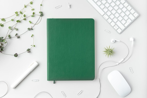 Flatlay of home office desk table Top view of workspace with green notebook keyboard mouse marker headphones pins and plants on white background