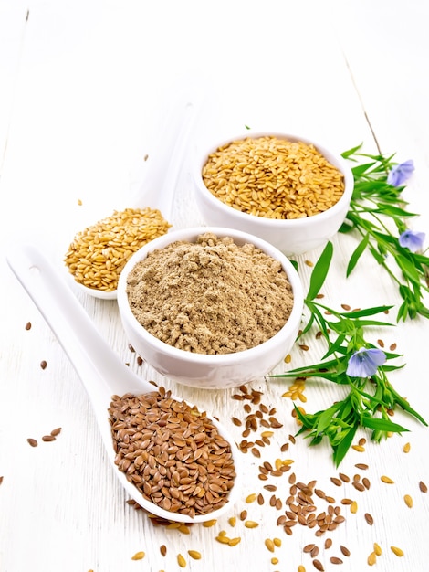 Flaxseed flour in a bowl, white and brown linen seeds in two spoons and on table, leaves and blue flax flowers on white wooden board background