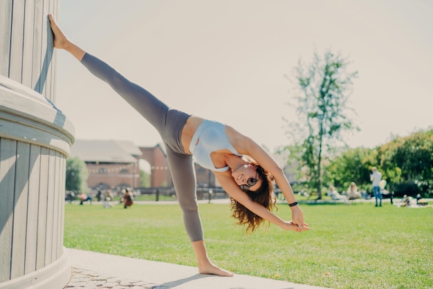 Flexible slim woman does stretching exercises outdoor stays in good physical shape dressed in cropped top and leggings stands on one leg leans with arms raised warms up before cardio training