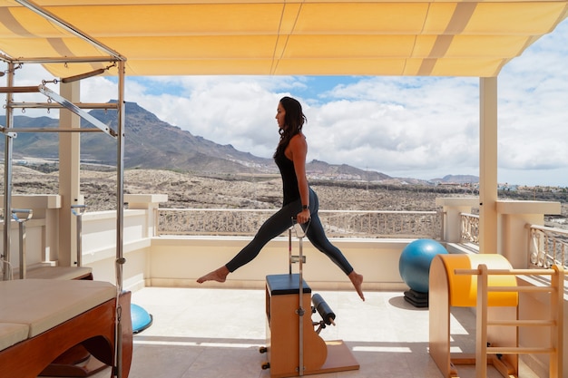 Flexible woman practicing pilates on summer day on special equipment, young fitness lady stretching legs with reformer while exercising on terrace outdoors with amazing mountains view. Sport concept