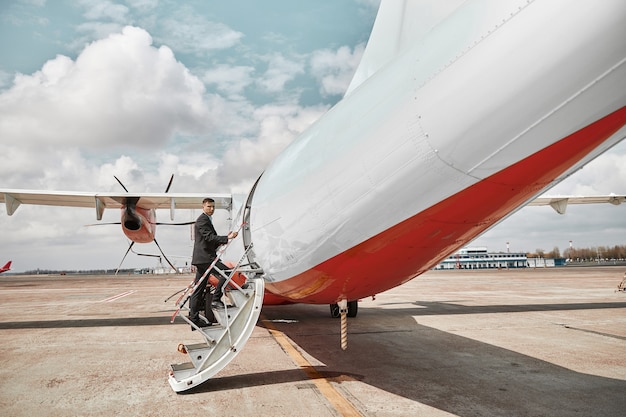 Flight attendant going up on ladder to airplane jet. Modern passenger plane. Serious mixed race man wear uniform. Sunny daytime. Civil commercial aviation. Air travel concept