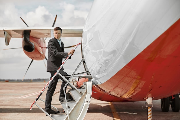 Flight attendant going up on ladder to airplane jet. Modern passenger plane. Serious multiracial man wear uniform. Sunny daytime. Civil commercial aviation. Air travel concept