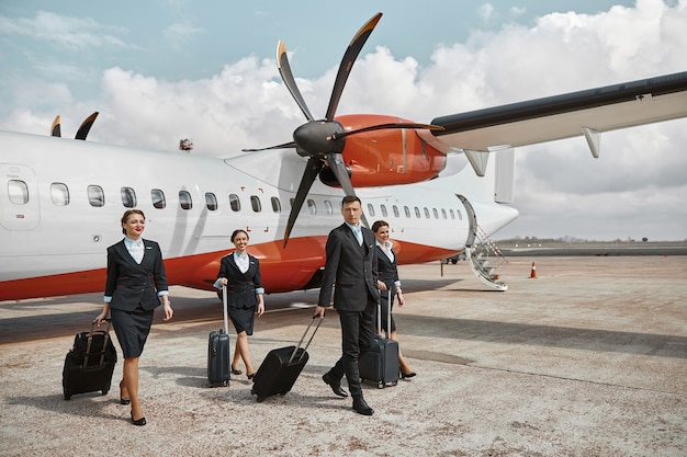 Flight attendant and stewardesses walking on runway near airplane jet. Modern passenger plane. Smiling man and women with baggage wear uniform. Teamwork. Civil commercial aviation. Air travel concept