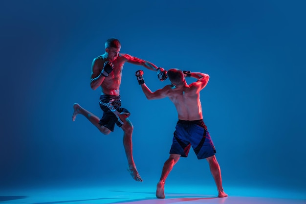 In flight. MMA. Two professional fighters punching or boxing isolated on blue studio background in neon. Fit muscular caucasian athletes or boxers fighting. Sport, competition and human emotions, ad.