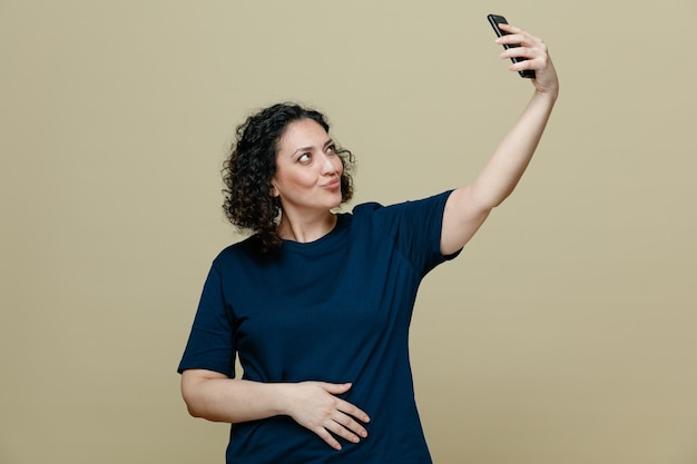 Flirty middleaged woman wearing tshirt keeping hand on belly raising mobile phone up making kiss gesture taking selfie isolated on olive green background