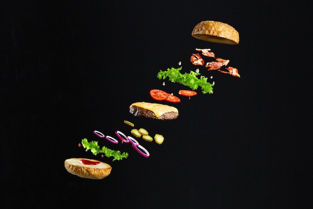 Floating burger isolated on black wooden background. Ingredients of a delicious burger with ground beef patty, lettuce, bacon, onions, tomatoes and cucumbers