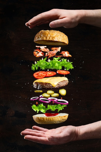 Floating burger isolated on black wooden background in men hands. Ingredients of a delicious burger with ground beef patty, lettuce, bacon, onions, tomatoes and cucumbers