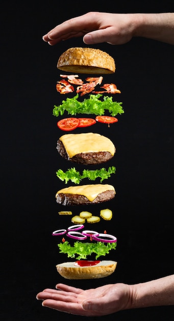 Floating burger isolated on black wooden background in men hands. Ingredients of a delicious burger with ground beef patty, lettuce, bacon, onions, tomatoes and cucumbers