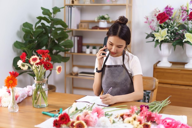 Florist concept Female florist talking with customer on phone and taking note in notebook at shop