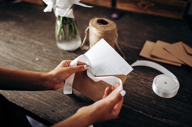 Florist is packing a postcard in an envelope with white ribbon on the wooden table .