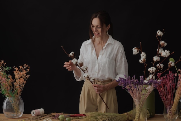 Florist makes a bouquet of dried flowers at a wooden table on a plain dark background with copyspace