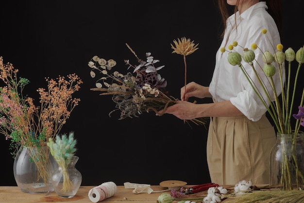 Florist makes a bouquet of dried flowers at a wooden table on a plain dark background with copyspace