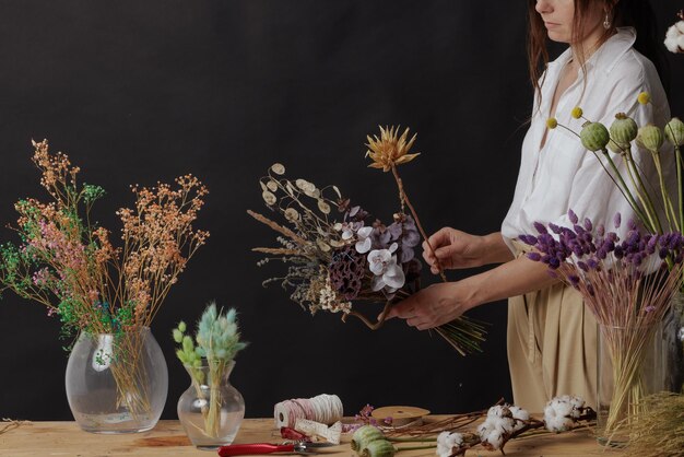 Florist makes a bouquet of dried flowers at a wooden table on a plain dark background with copyspace