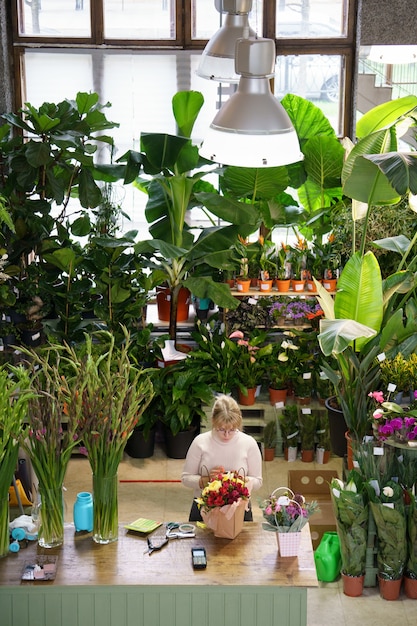 Florist at workplace flower shop owner creating bouquet of roses with houseplants on racks behind