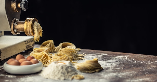 Flour, eggs and a pasta maker producing fresh Italian fettuccine with a dark background.